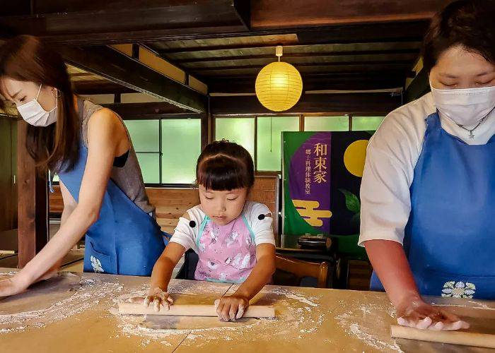 Three people rolling out udon dough in a traditional Kyoto townhouse; in the center is a young girl.